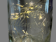 Das Bild zeigt eine elegante Glasflasche mit warmweiß leuchtender LED‑Lichterkette im Inneren. Auf der Flasche ist der Schriftzug „Frohe Weihnachten“ eingraviert, der durch das Licht sanft hervorgehoben wird